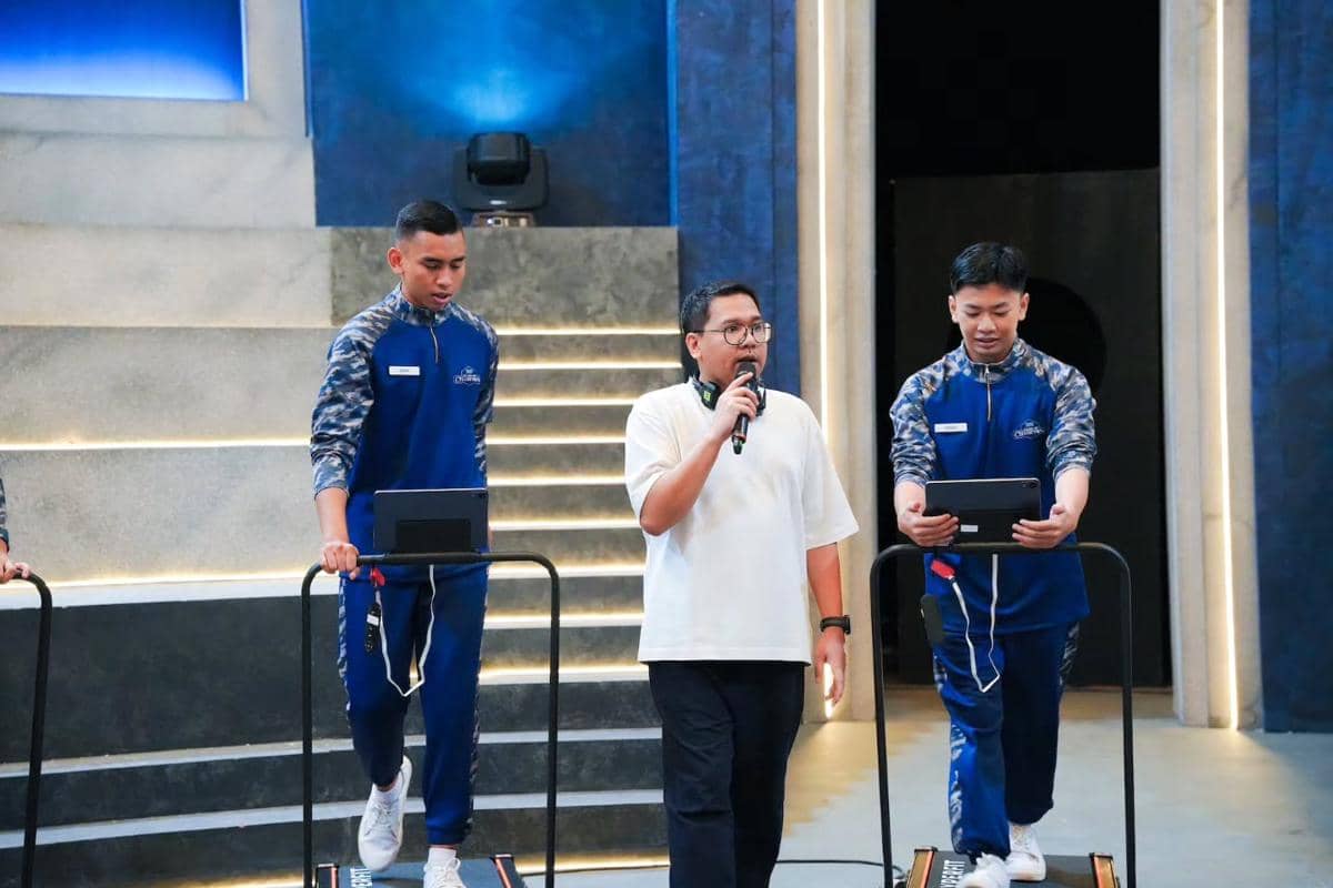 Dua peserta mengenakan seragam biru berlari di atas treadmill sambil menjawab soal, sementara seorang pria memegang mikrofon di tengah mereka.