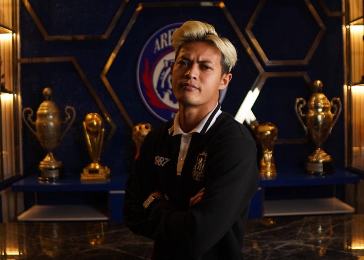 Seorang pria berambut pirang mengenakan jaket hitam berpose dengan tangan terlipat di depan logo Arema FC dan deretan trofi emas.