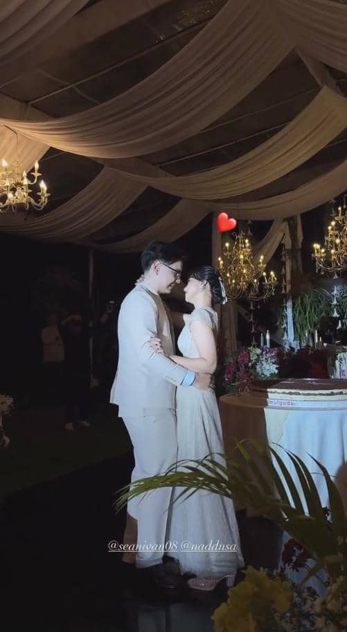 Sepasang pengantin mengenakan busana formal berwarna terang sedang berdansa di bawah dekorasi tenda elegan dengan lampu gantung mewah.