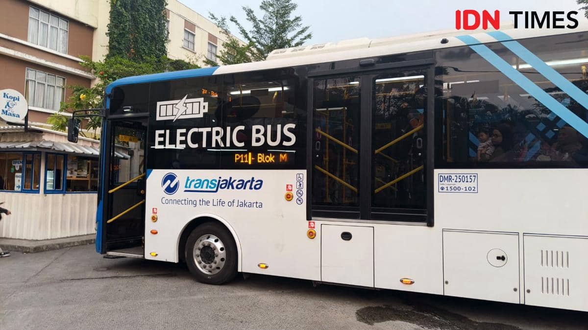 Bus listrik Transjakarta rute P11 Bogor-Blok M berhenti di tepi jalan dengan penumpang di dalamnya dan bangunan di latar belakang.