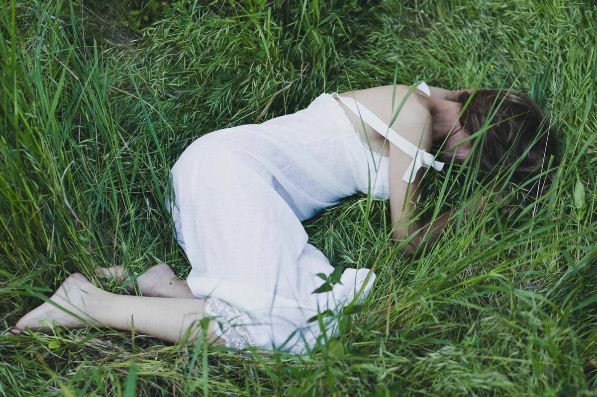Seorang perempuan mengenakan gaun putih terbaring di rerumputan hijau dengan posisi meringkuk di alam terbuka.
