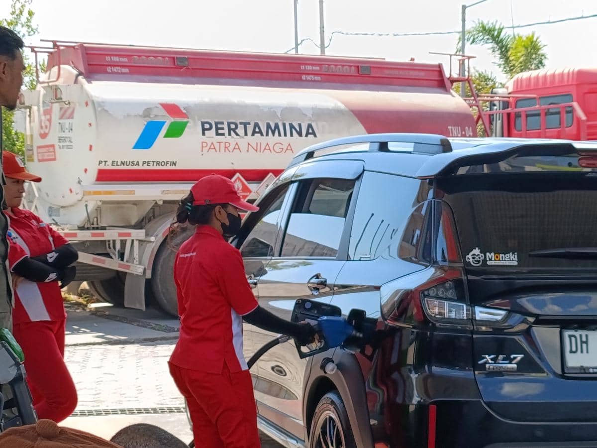 Petugas SPBU mengenakan seragam merah sedang mengisi BBM Pertamax ke mobil hitam di Kota Kupang dengan truk tangki Pertamina di latar belakang.