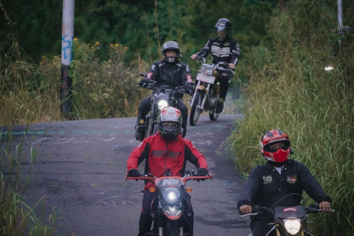 Beberapa pengendara motor dengan perlengkapan lengkap melintasi jalan pedesaan dikelilingi pepohonan dan rerumputan tinggi.