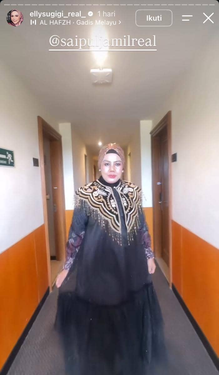 Seorang perempuan mengenakan gaun hitam berhias bordir emas dan hijab cokelat berjalan di lorong hotel dengan pencahayaan hangat.