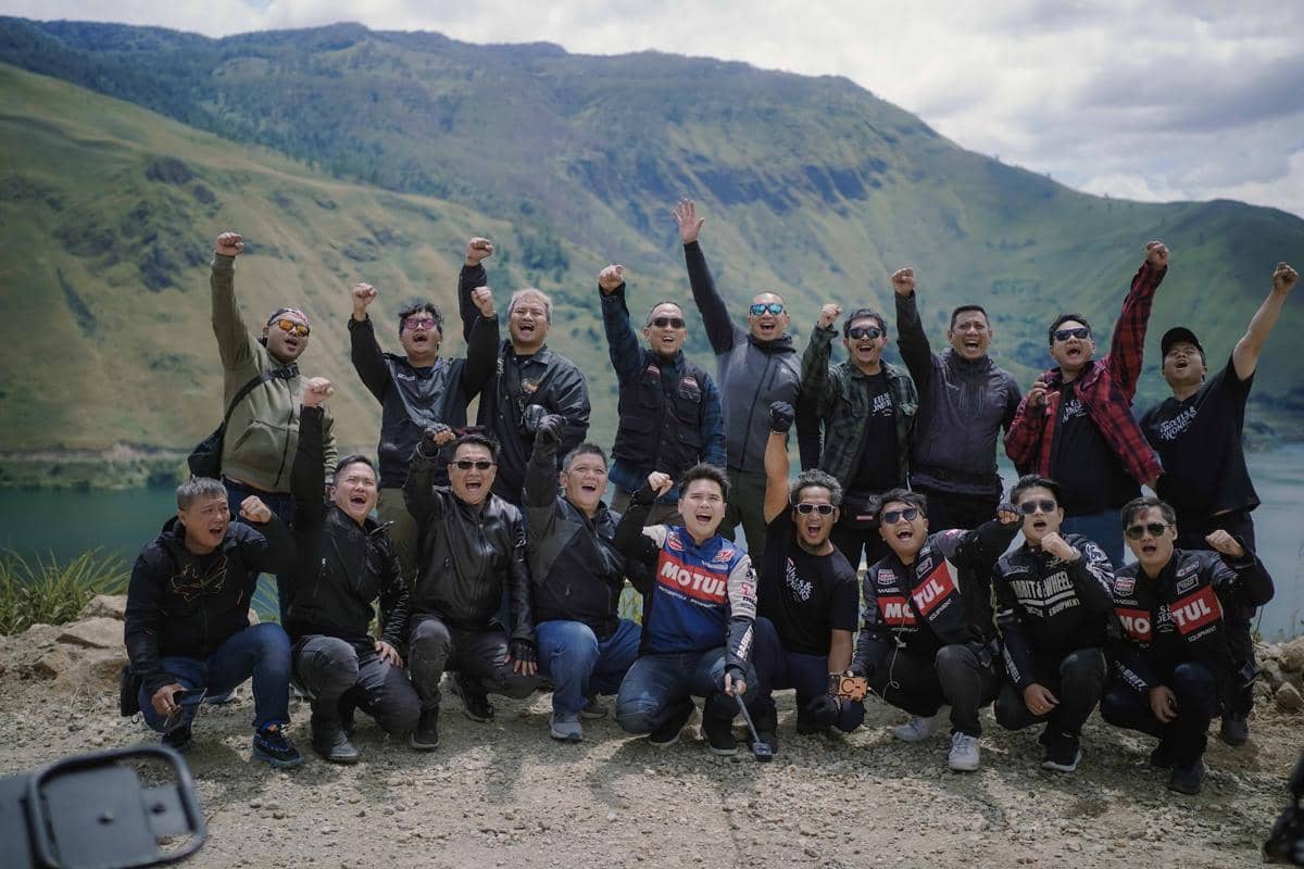 Sekelompok bikers mengenakan jaket dan kacamata hitam berpose gembira di tepi Danau Toba dengan latar pegunungan hijau di Samosir.