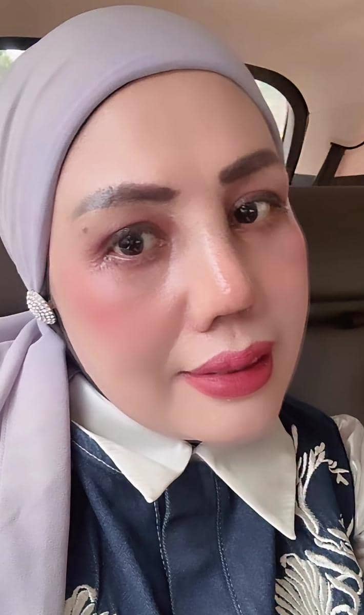 Seorang perempuan mengenakan hijab abu-abu muda dan atasan berkerah putih dengan bordir bunga, berpose di dalam mobil.