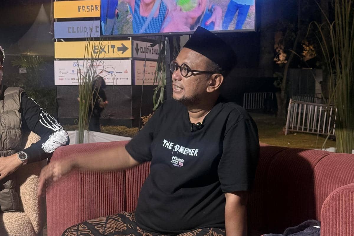 Seorang pria mengenakan kaus hitam dan peci duduk di sofa merah saat wawancara di area Prambanan Jazz Festival 2026 pada malam hari.