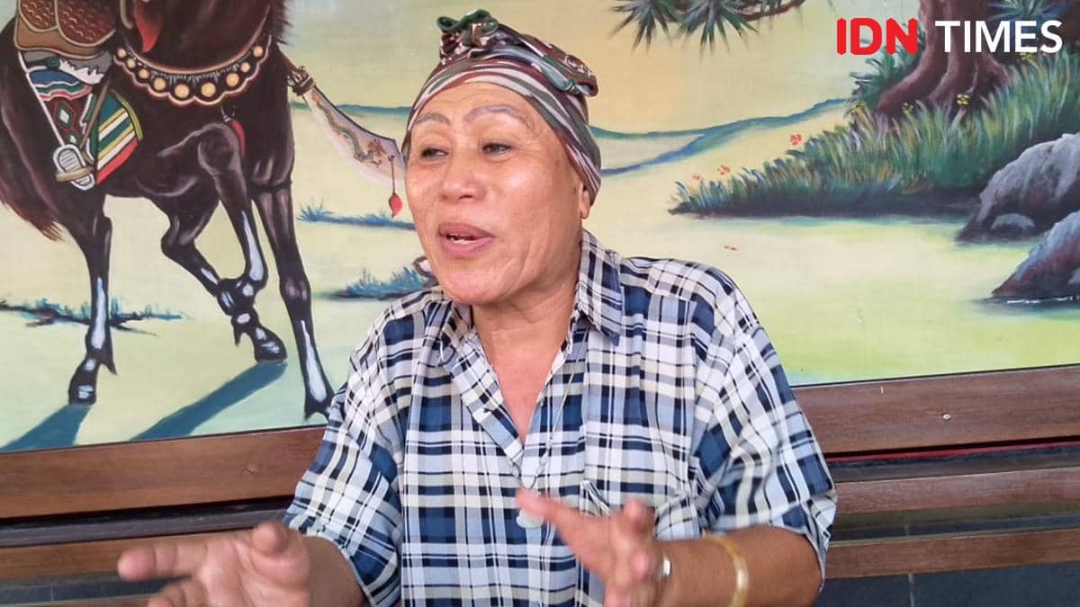 Seorang perempuan mengenakan kemeja kotak-kotak dan penutup kepala bermotif sedang berbicara dengan ekspresif di depan lukisan kuda.