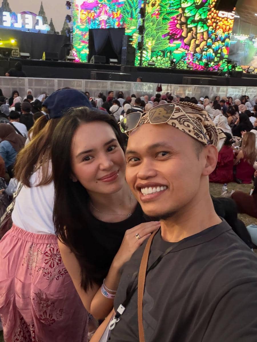 Dua orang berpose sambil tersenyum di area penonton Prambanan Jazz Festival 2026 dengan panggung berwarna cerah di latar belakang.
