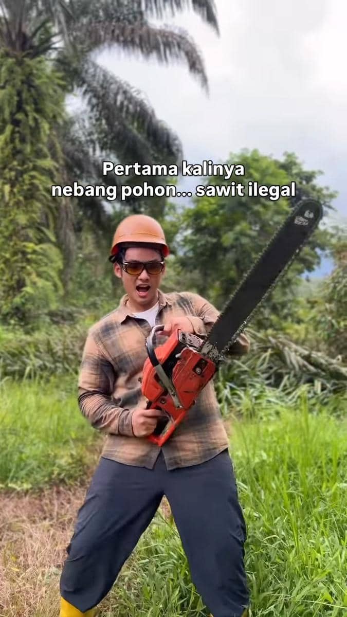 Seorang pria mengenakan helm keselamatan dan kacamata pelindung memegang gergaji mesin di area perkebunan sawit dengan ekspresi bersemangat.