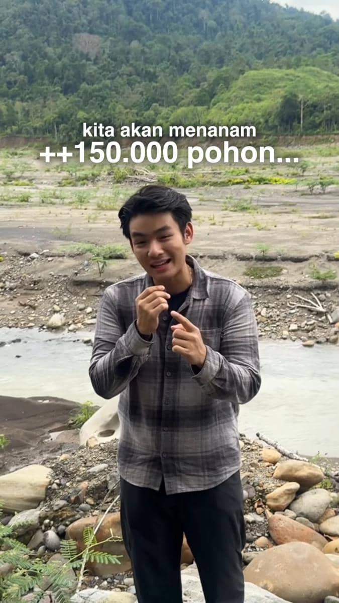 Seorang pria berbicara di tepi sungai dengan latar belakang perbukitan hijau dan teks bertuliskan 'kita akan menanam ++150.000 pohon'.