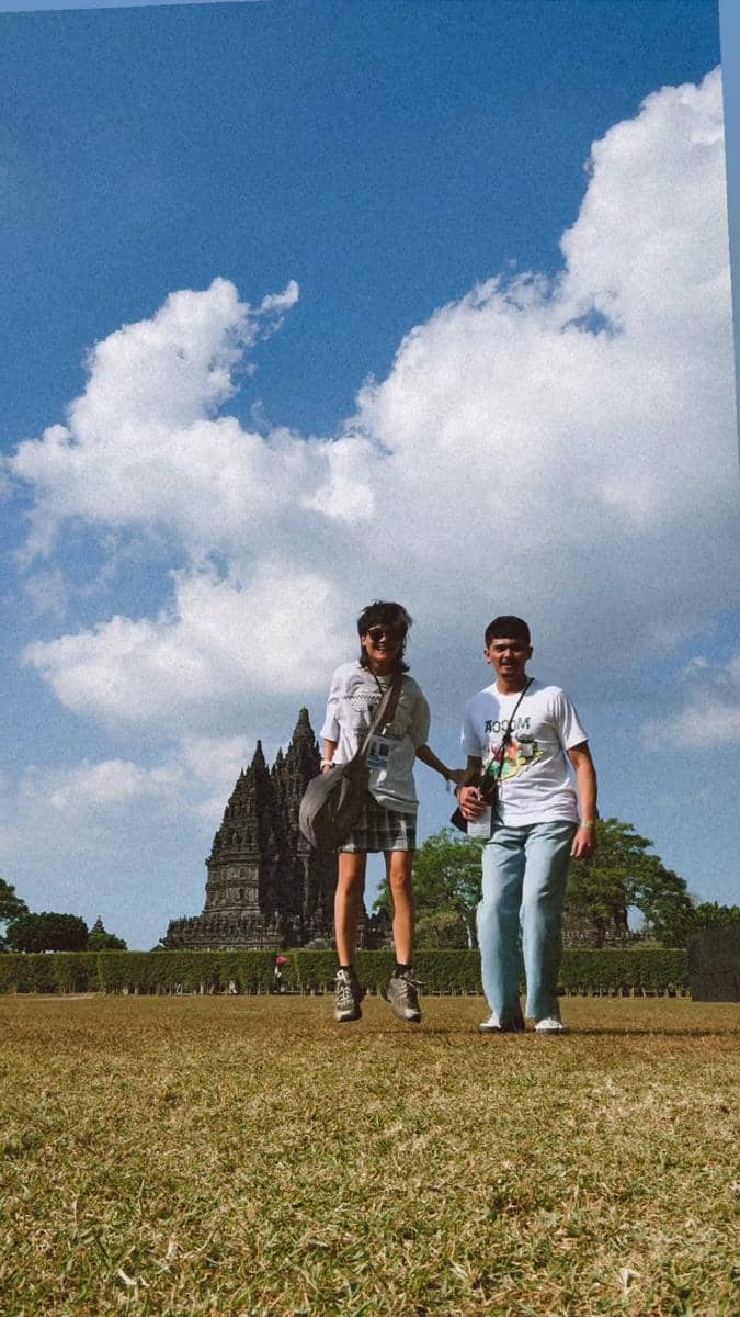 Dua orang berpose ceria di area Candi Prambanan dengan langit biru dan awan putih di latar belakang saat festival musik.