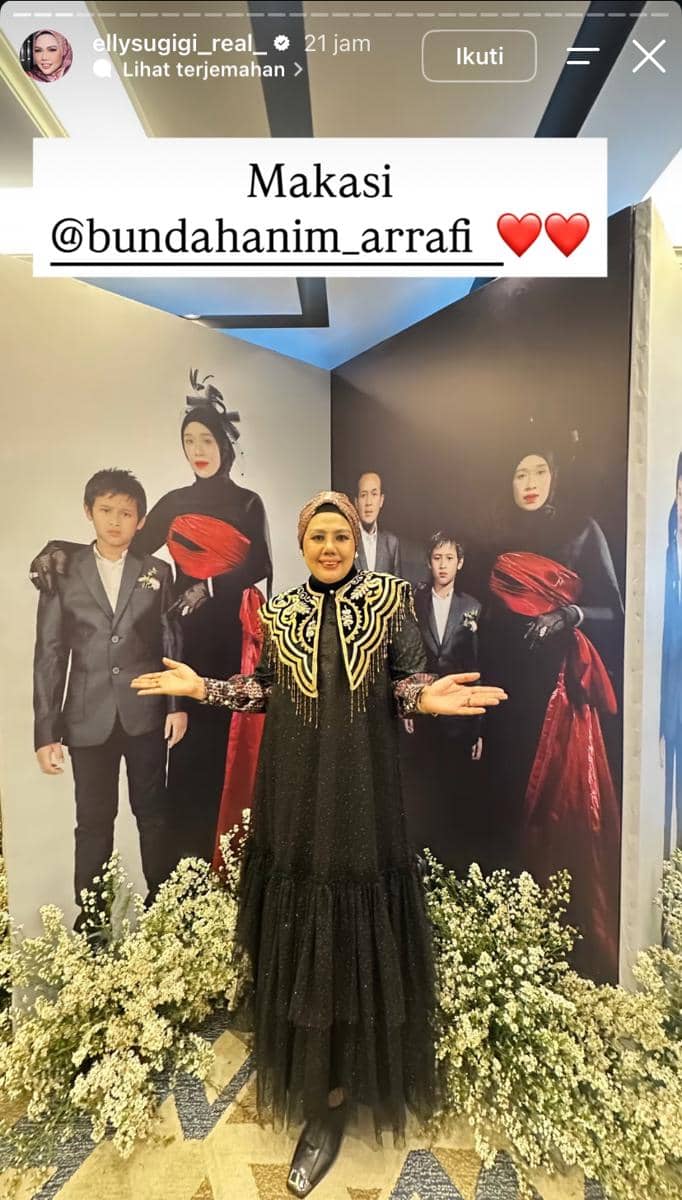 Seorang perempuan mengenakan gaun hitam berhias emas berpose di depan latar foto keluarga dengan dekorasi bunga putih di sekitarnya.