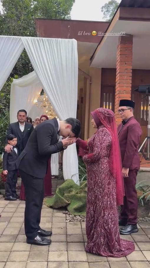 Seorang pria bersetelan jas hitam mencium tangan wanita berhijab merah marun dalam upacara pernikahan di luar ruangan.