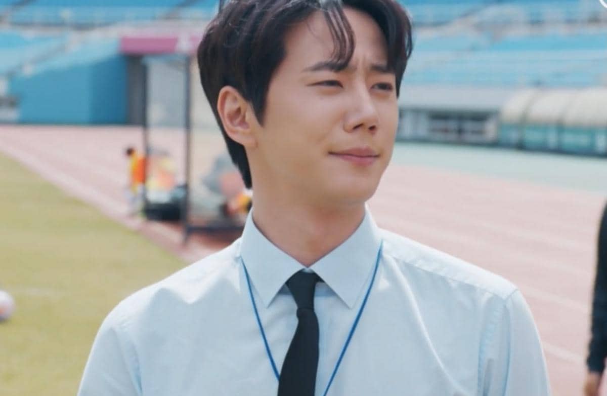 Seorang pria mengenakan kemeja biru muda dan dasi hitam berdiri di tepi lapangan olahraga dalam cuplikan drama Korea Reborn Rookie.