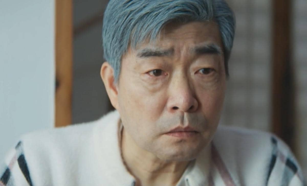 Seorang pria berambut abu-abu mengenakan sweter putih bermotif garis terlihat serius dalam cuplikan drama Korea Reborn Rookie.