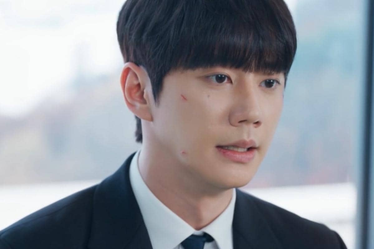 Seorang pria mengenakan setelan jas hitam dan dasi biru tampak serius dengan beberapa luka kecil di wajahnya dalam cuplikan drama Reborn Rookie.