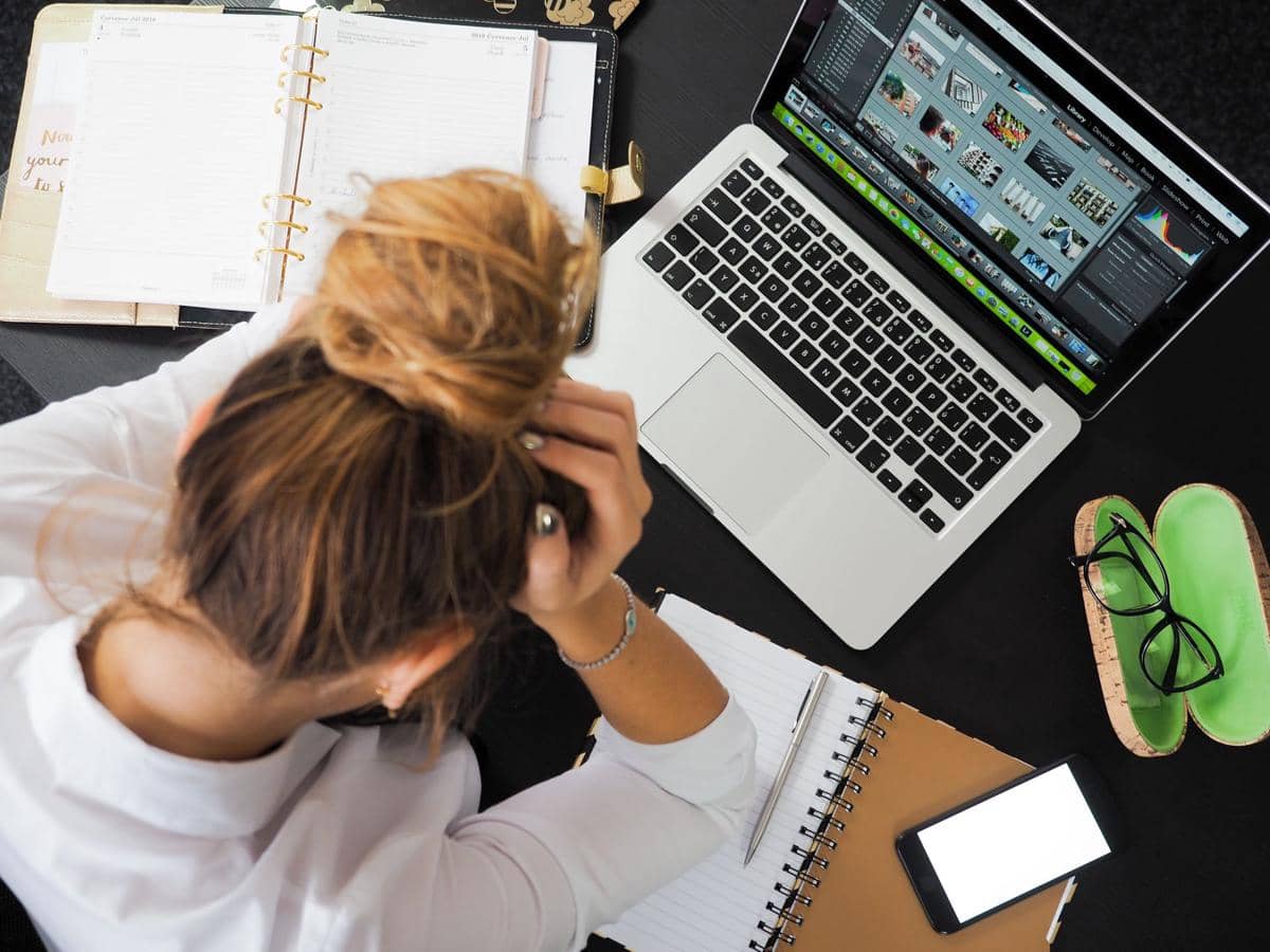 Seorang perempuan memegang kepala di depan laptop dan buku catatan di meja kerja, tampak lelah saat bekerja di kantor.