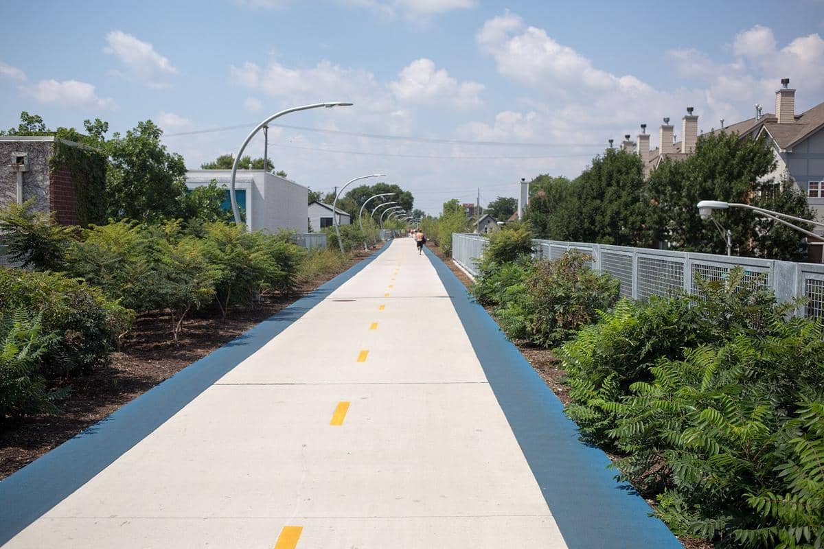 Jalur pejalan kaki dan sepeda The 606 Trail dengan marka kuning di tengah, dikelilingi pepohonan dan bangunan di bawah langit cerah.