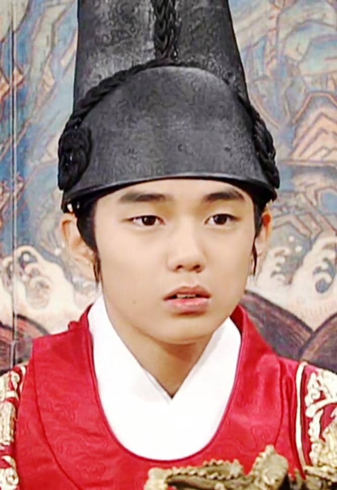 Seorang aktor muda mengenakan hanbok kerajaan berwarna merah dan topi tradisional hitam dalam adegan drama sejarah Korea.