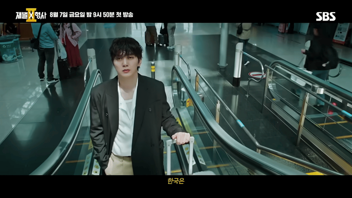 Seorang pria mengenakan jas hitam dan kemeja putih berdiri di eskalator dalam cuplikan teaser drama Flex x Cop 2 dari SBS.