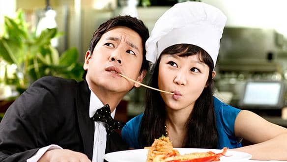 Dua orang sedang berbagi seutas spaghetti di dapur, pria mengenakan setelan jas dan wanita memakai seragam koki dengan topi putih.