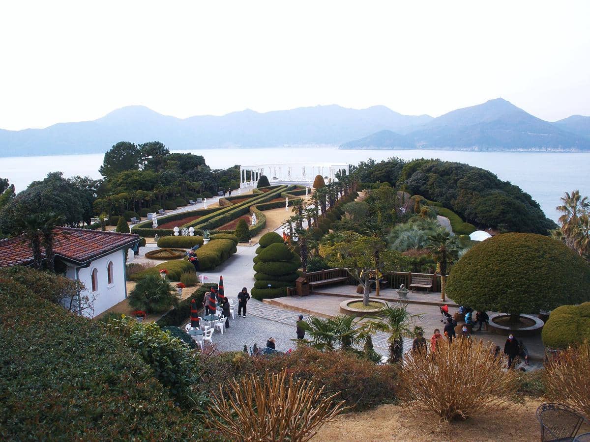 Pemandangan taman indah di Pulau Oedo Botania dengan pepohonan rapi, jalur pejalan kaki, dan laut biru di latar belakang pegunungan.