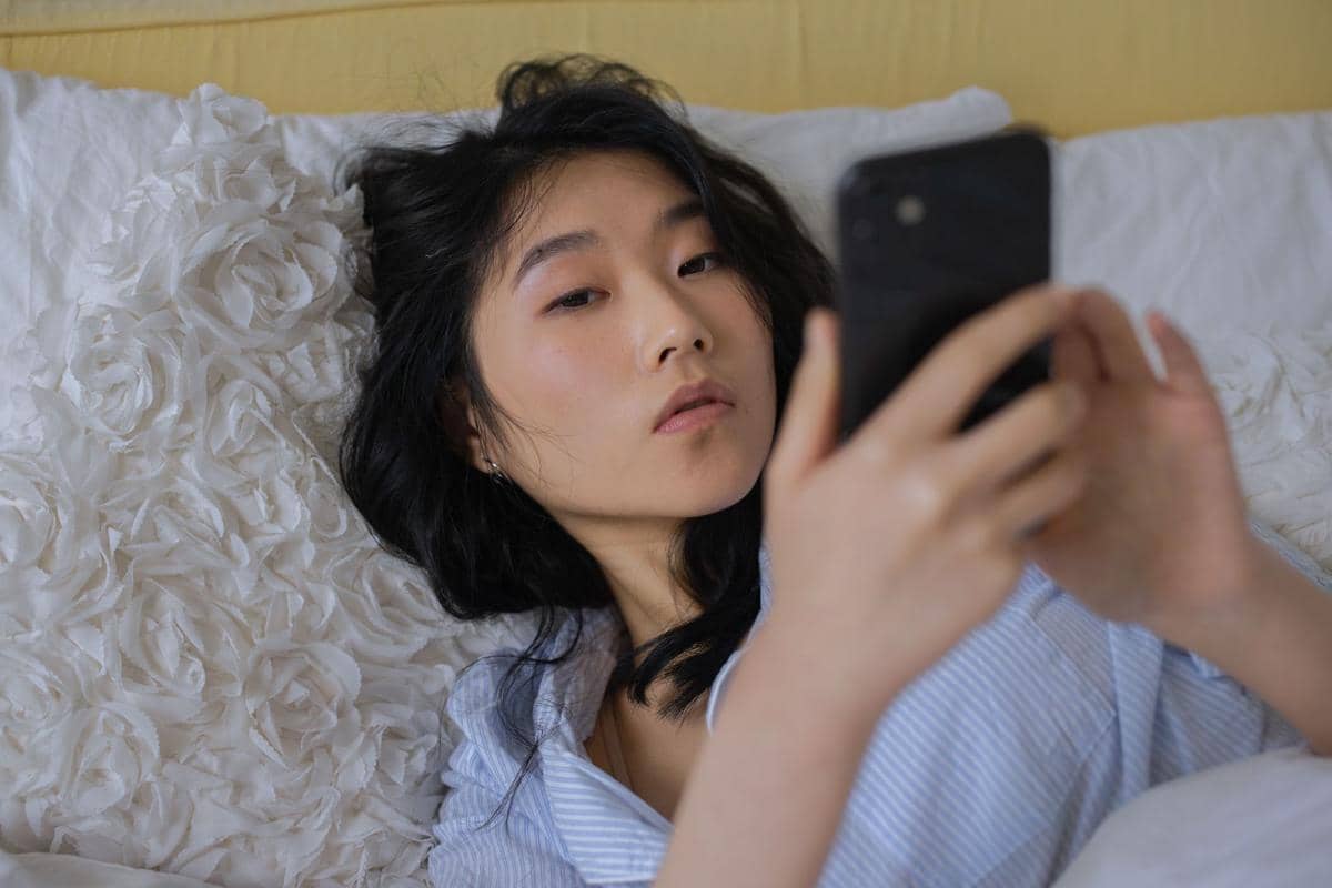 Perempuan berbaring di tempat tidur sambil menggunakan ponsel.