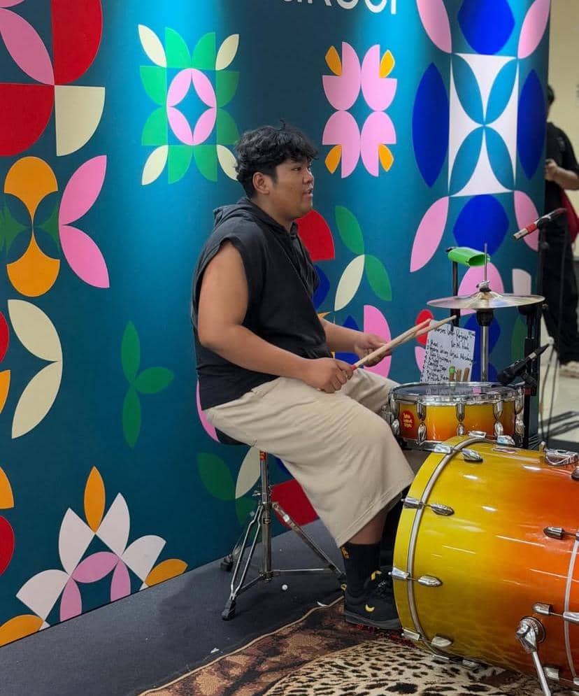 Seorang pemain drum duduk di belakang set drum berwarna oranye gradasi, tampil di depan dinding bermotif warna-warni dengan pola geometris.