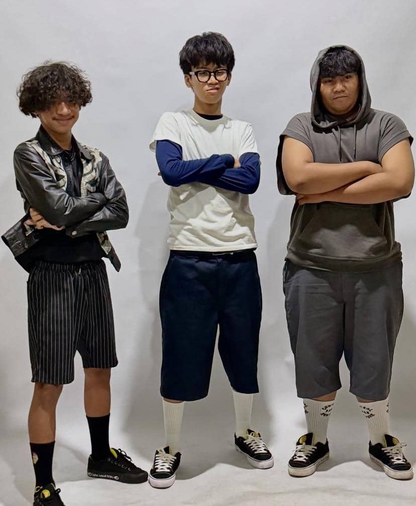 Tiga anggota grup musik berdiri berpose dengan tangan bersilang di depan latar putih, mengenakan pakaian kasual dan sepatu kets.