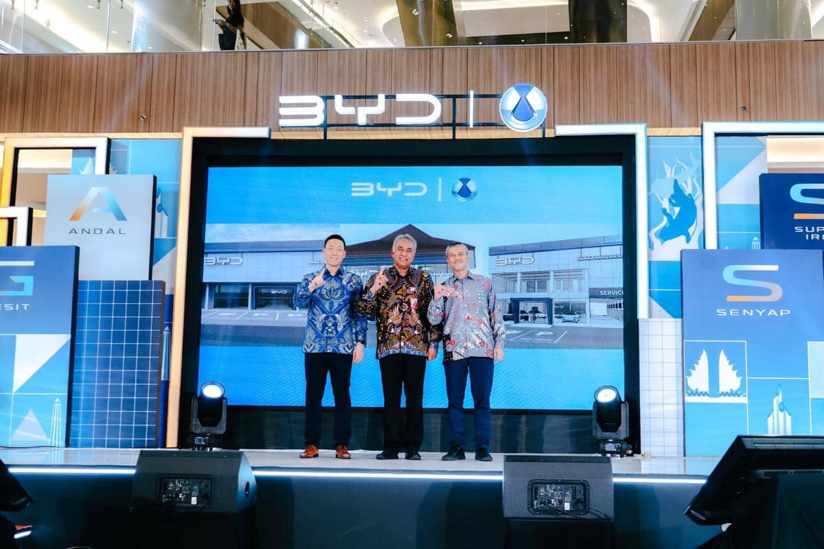 Tiga pria berbatik berdiri di panggung acara perluasan diler BYD di Jawa Timur dengan latar layar besar bergambar gedung diler.