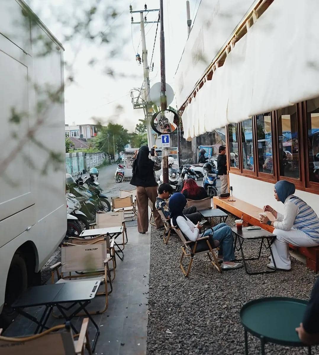Suasana outdoor di Cafe Fatboy Dining Club dengan beberapa pengunjung duduk santai di kursi kayu di tepi jalan berkerikil.