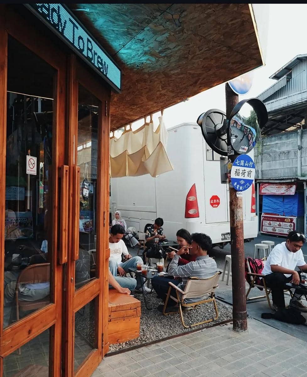 Beberapa orang duduk santai di area outdoor Cafe Fatboy Dining Club dengan minuman di meja dan suasana sore yang hangat.