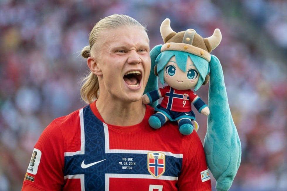 Seorang pemain sepak bola mengenakan jersey Norwegia berteriak dengan boneka karakter anime berhelm bertanduk di pundaknya di stadion.