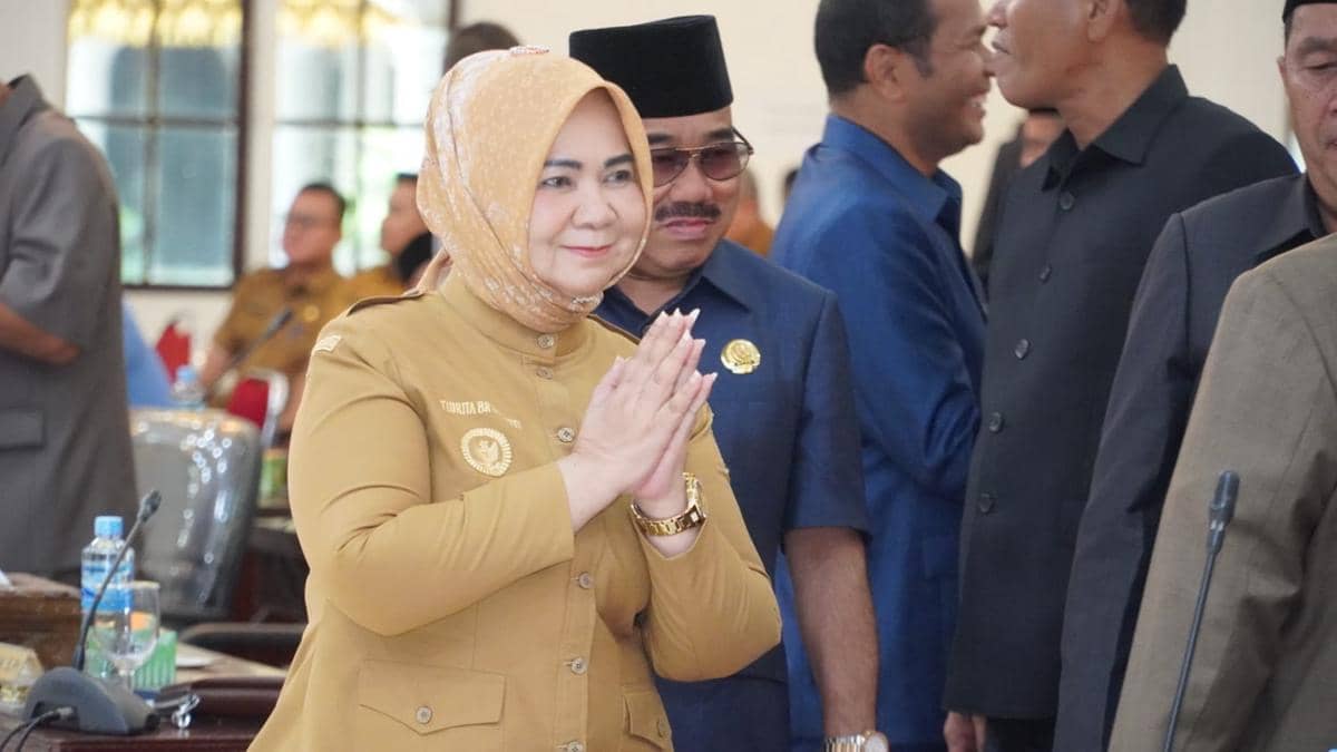 Seorang perempuan berjilbab mengenakan seragam cokelat melakukan salam dengan tangan di dada dalam sebuah acara resmi di dalam ruangan.