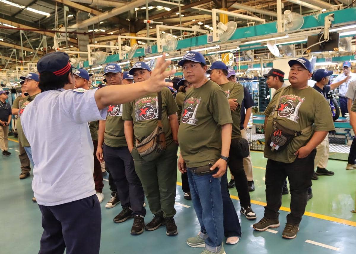 Seorang pemandu menjelaskan proses produksi motor Yamaha kepada sekelompok peserta yang mengenakan kaus hijau dan topi biru di area pabrik.