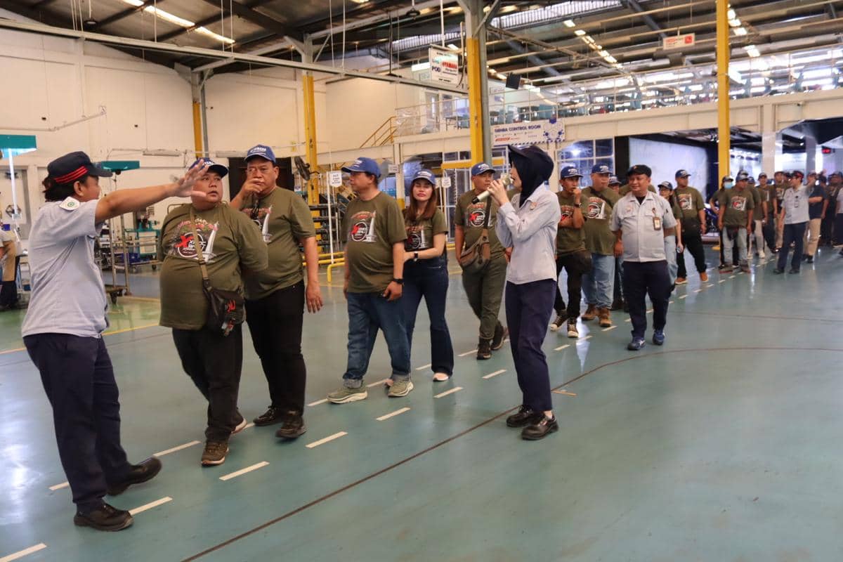 Sejumlah peserta mengenakan kaus hijau dan topi biru mengikuti tur pabrik Yamaha yang dipandu oleh petugas di area produksi.
