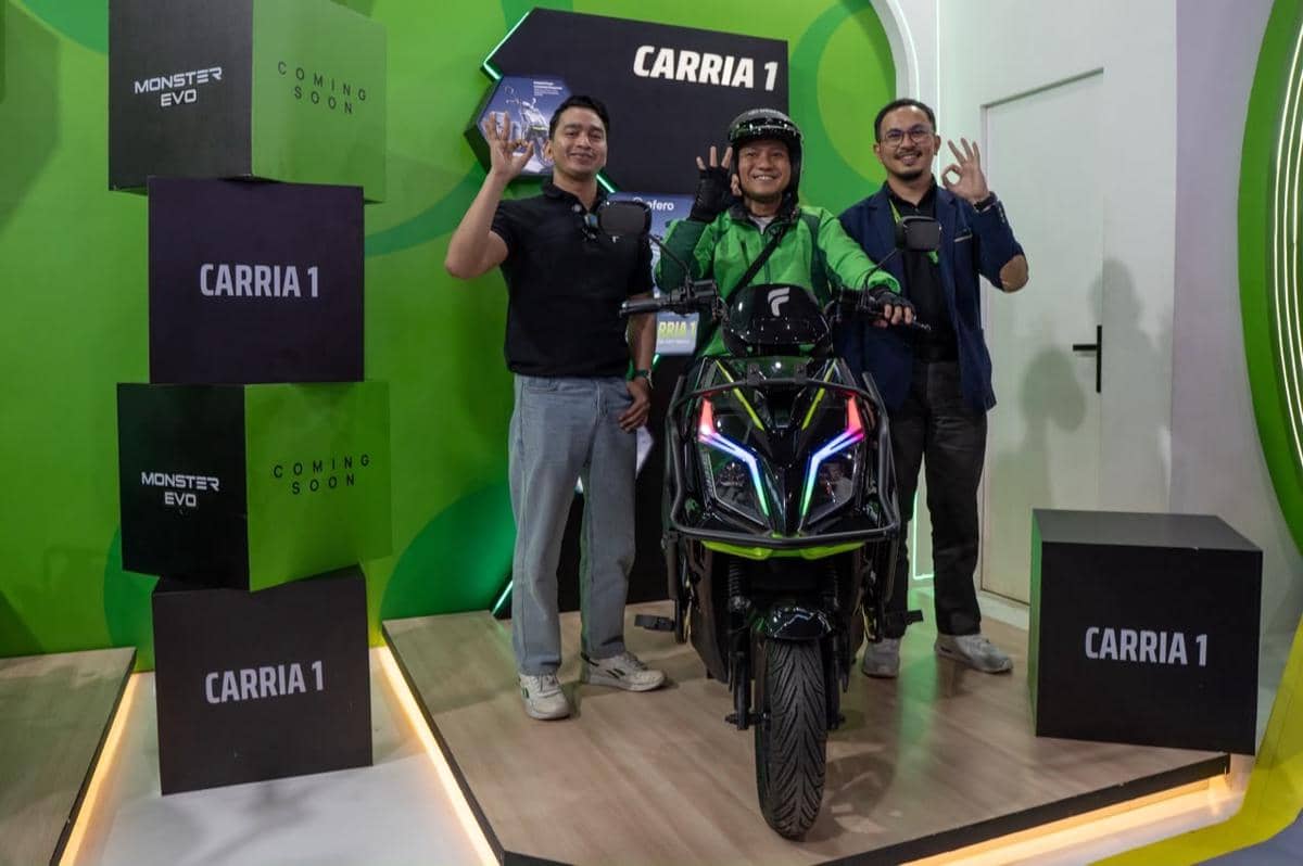 Tiga pria berpose di samping motor listrik Ofero CarriA 1 berwarna hitam hijau di area pameran dengan latar hijau bertuliskan CarriA 1.