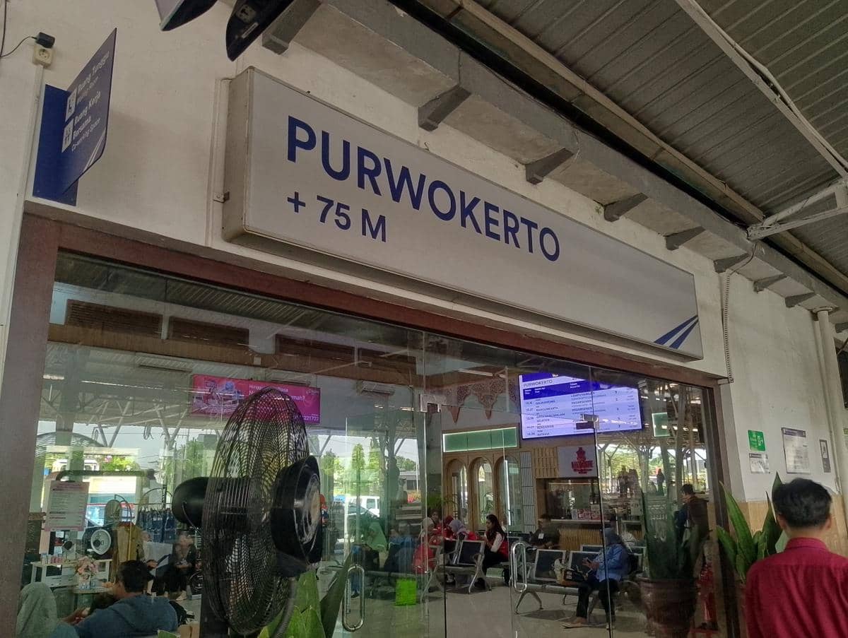 Area ruang tunggu Stasiun Purwokerto dengan papan nama bertuliskan Purwokerto dan penumpang yang duduk di dalam ruangan.