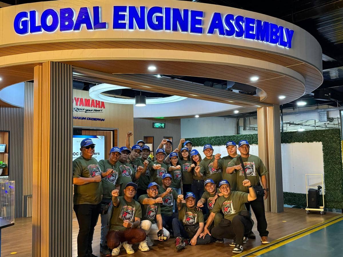 Anggota komunitas XMOC berfoto bersama di area Global Engine Assembly pabrik Yamaha dengan mengenakan kaus hijau dan topi biru.