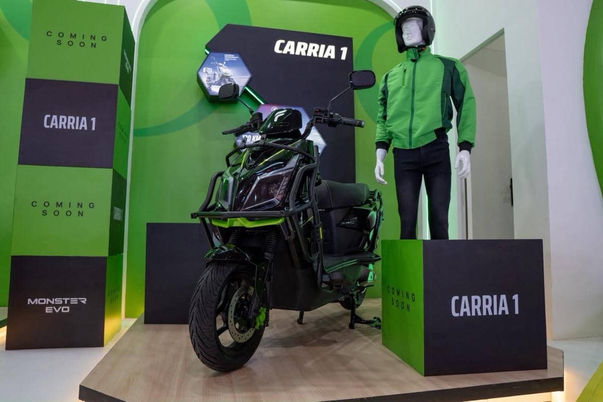 Motor listrik hitam hijau model CarriA 1 dipamerkan di booth dengan latar hijau bersama manekin berjaket hijau dan helm hitam.