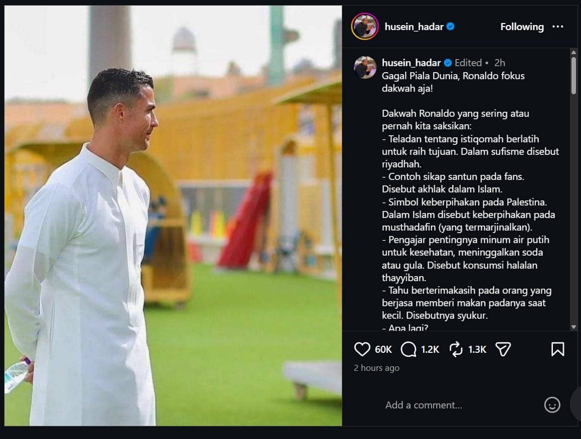 Seorang pria mengenakan jubah putih berdiri di area lapangan hijau dengan latar belakang bangunan berwarna kuning dan langit cerah.