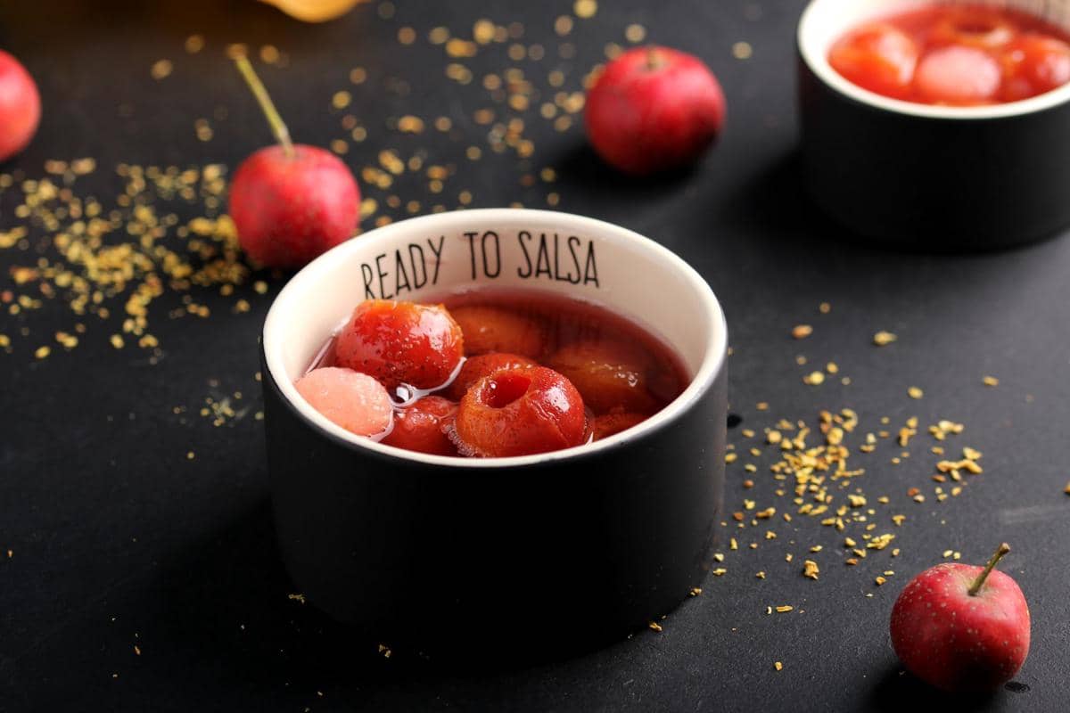 Semangkuk asinan ceri berwarna merah segar dalam wadah hitam bertuliskan 'Ready to Salsa' di atas meja gelap dengan taburan rempah.