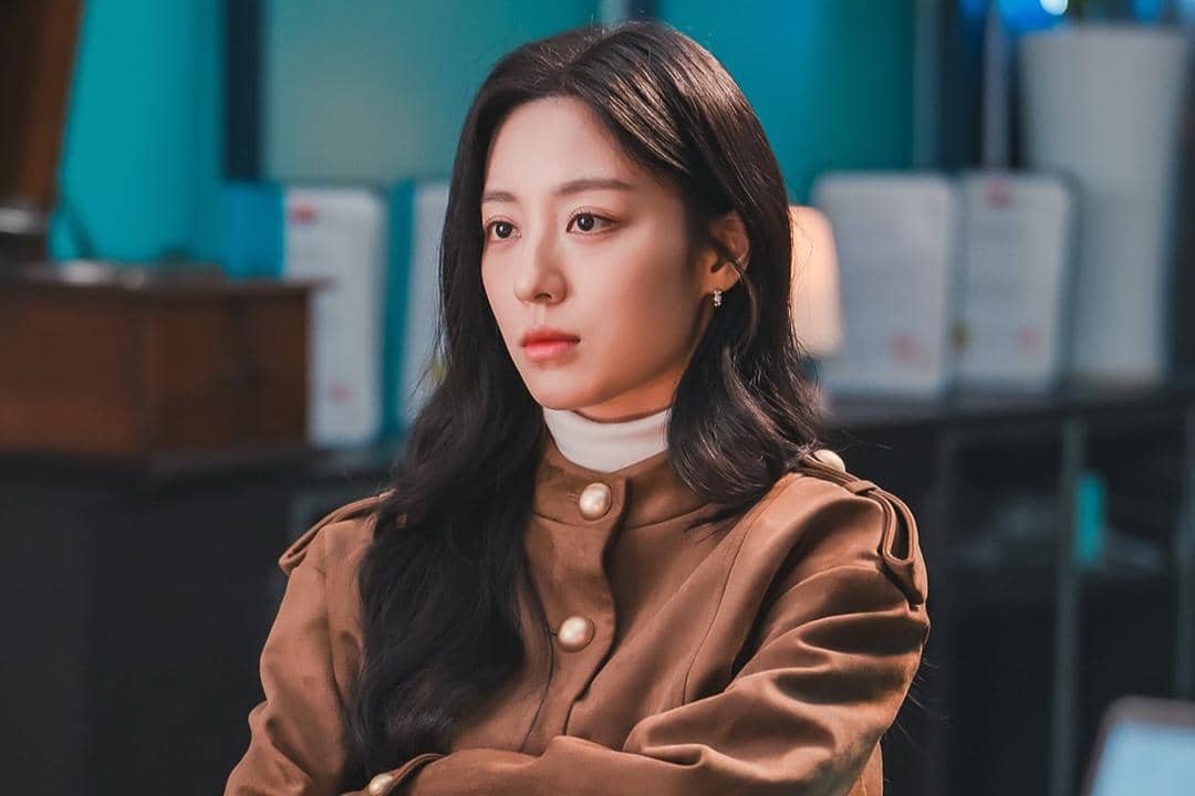 Seorang wanita berambut panjang mengenakan mantel cokelat duduk di ruangan dengan pencahayaan hangat dalam adegan drama Love In Sync.
