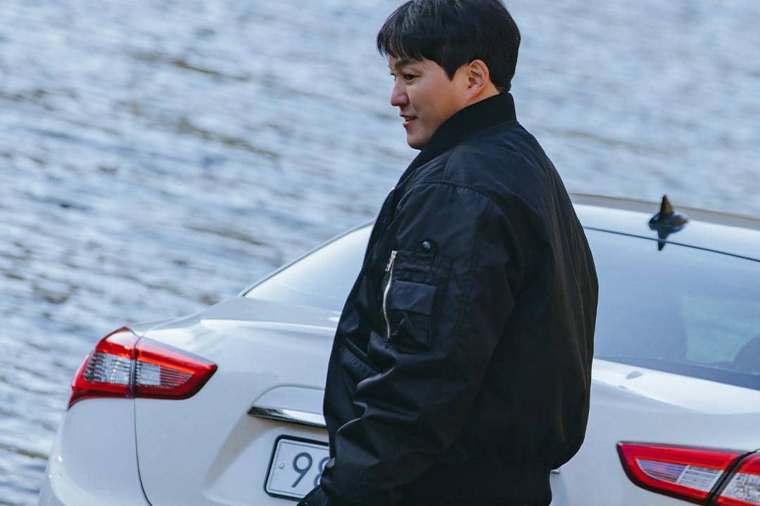 Seorang pria mengenakan jaket hitam berdiri di samping mobil putih di tepi perairan dalam adegan drama The Husband.