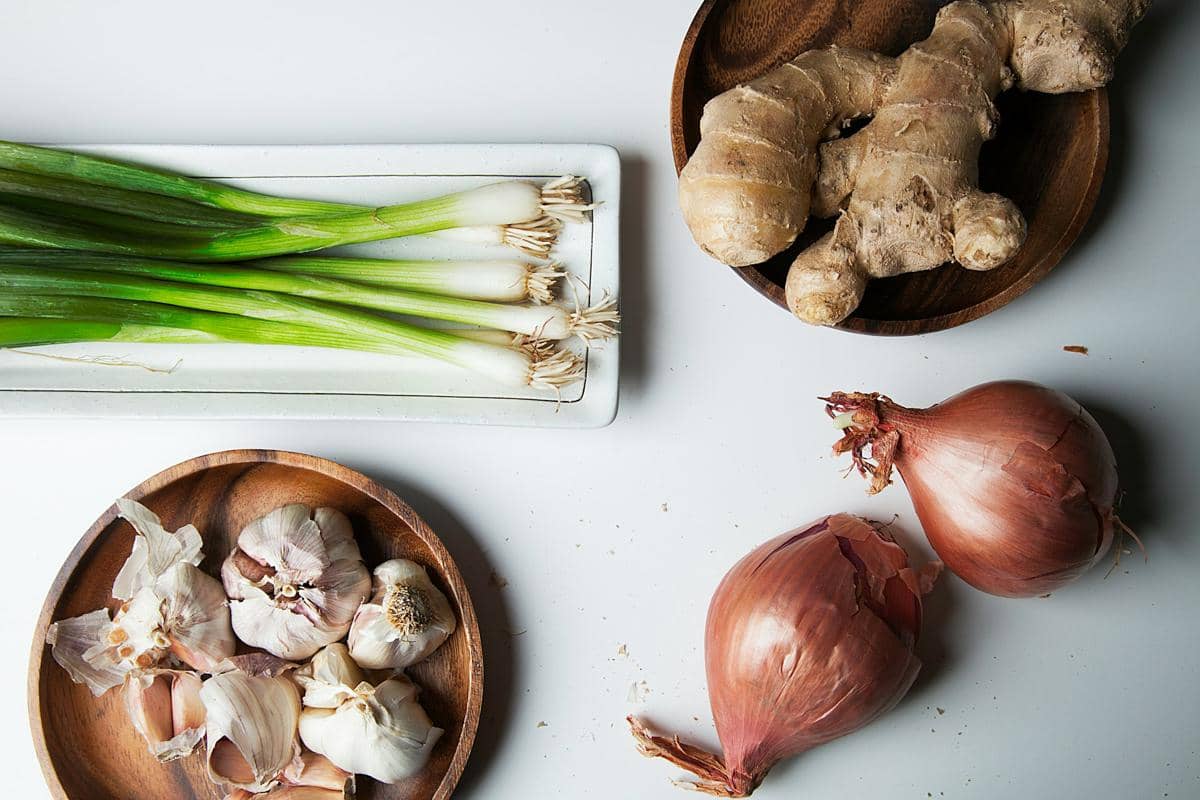 Beberapa bahan dapur seperti bawang putih, bawang merah, jahe, dan daun bawang tersusun di atas meja dengan wadah kayu.