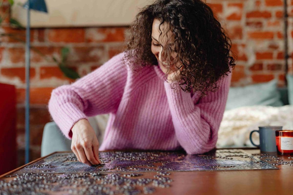 Seorang wanita mengenakan sweter ungu muda sedang merakit puzzle di atas meja kayu di ruangan dengan dinding bata merah.