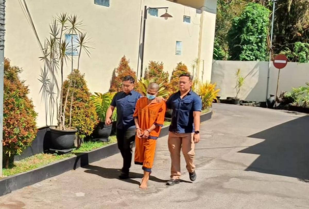 Seorang pria berpakaian tahanan oranye diborgol dan dikawal dua petugas berpakaian sipil menuju gedung Satreskrim Polres Malang.