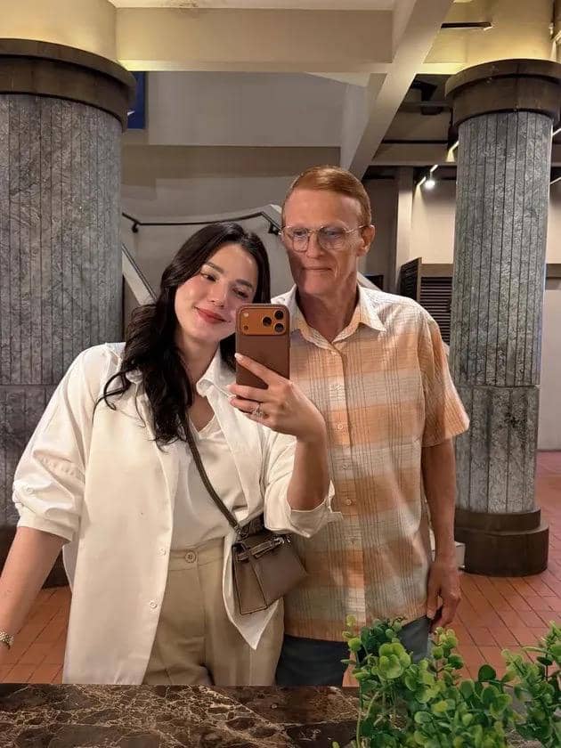 Seorang wanita berpose sambil berfoto cermin bersama seorang pria di ruangan dengan pilar besar dan pencahayaan hangat.