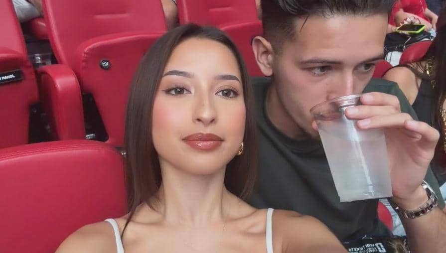 Seorang perempuan berpose untuk swafoto bersama seorang pria yang sedang memegang gelas minuman di tempat duduk stadion berwarna merah.